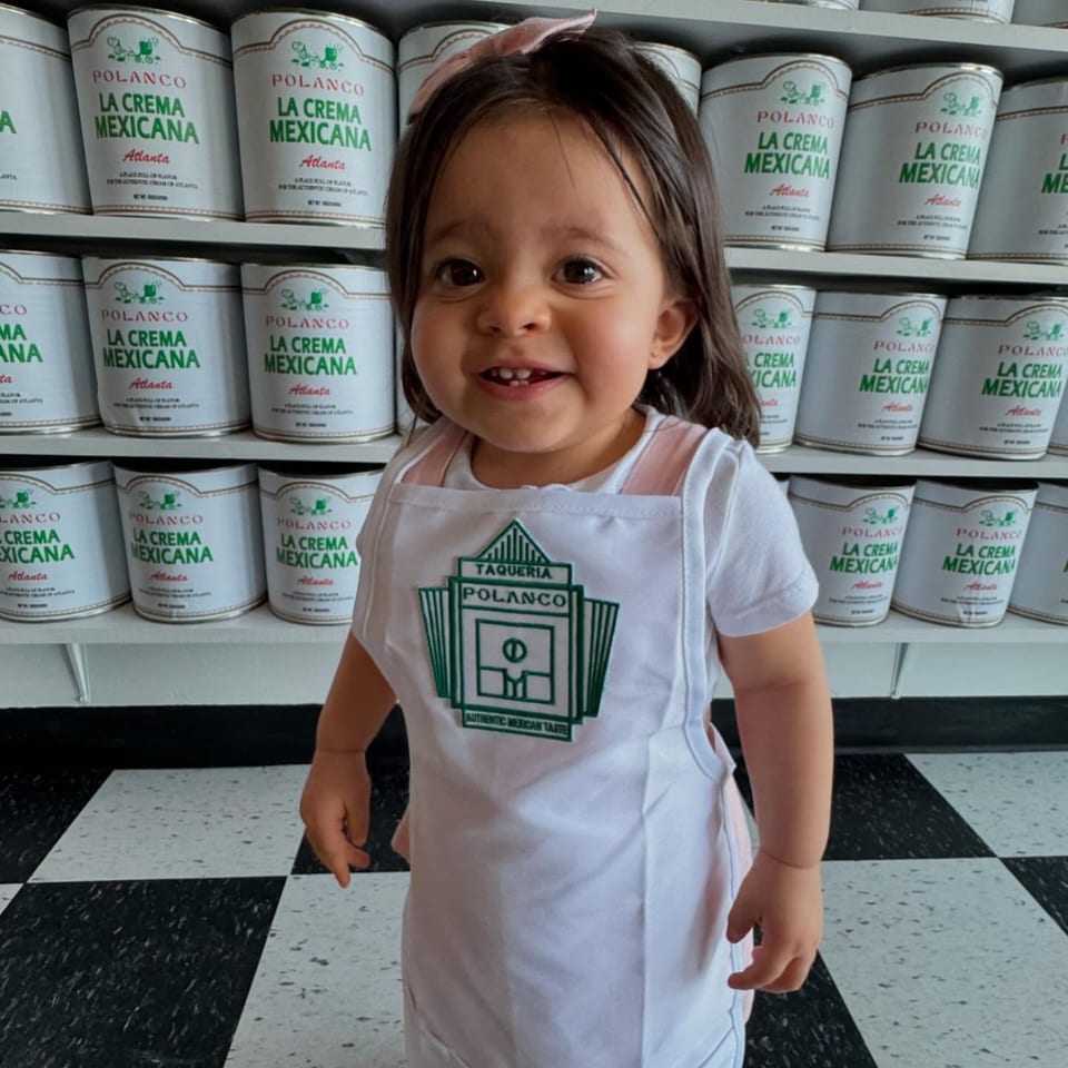 A smiling child in a Polanco Taqueria apron stands before shelves of "La Crema Mexicana" containers.