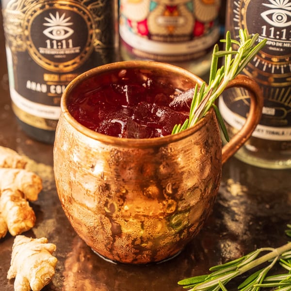 A copper mug filled with a red drink, ice, and rosemary, with ginger and bottles in the background.