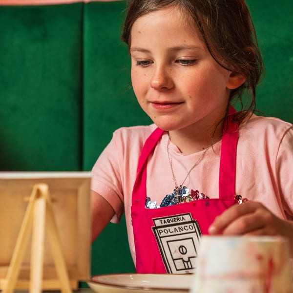 Young girl in a pink Polanco Taqueria apron painting, with a green background.
