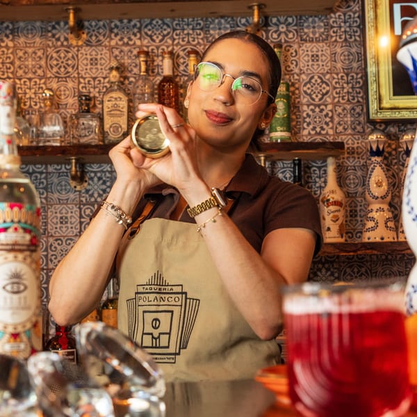 A Polanco Taqueria bartender in glasses and an apron shakes a cocktail shaker behind a bar.