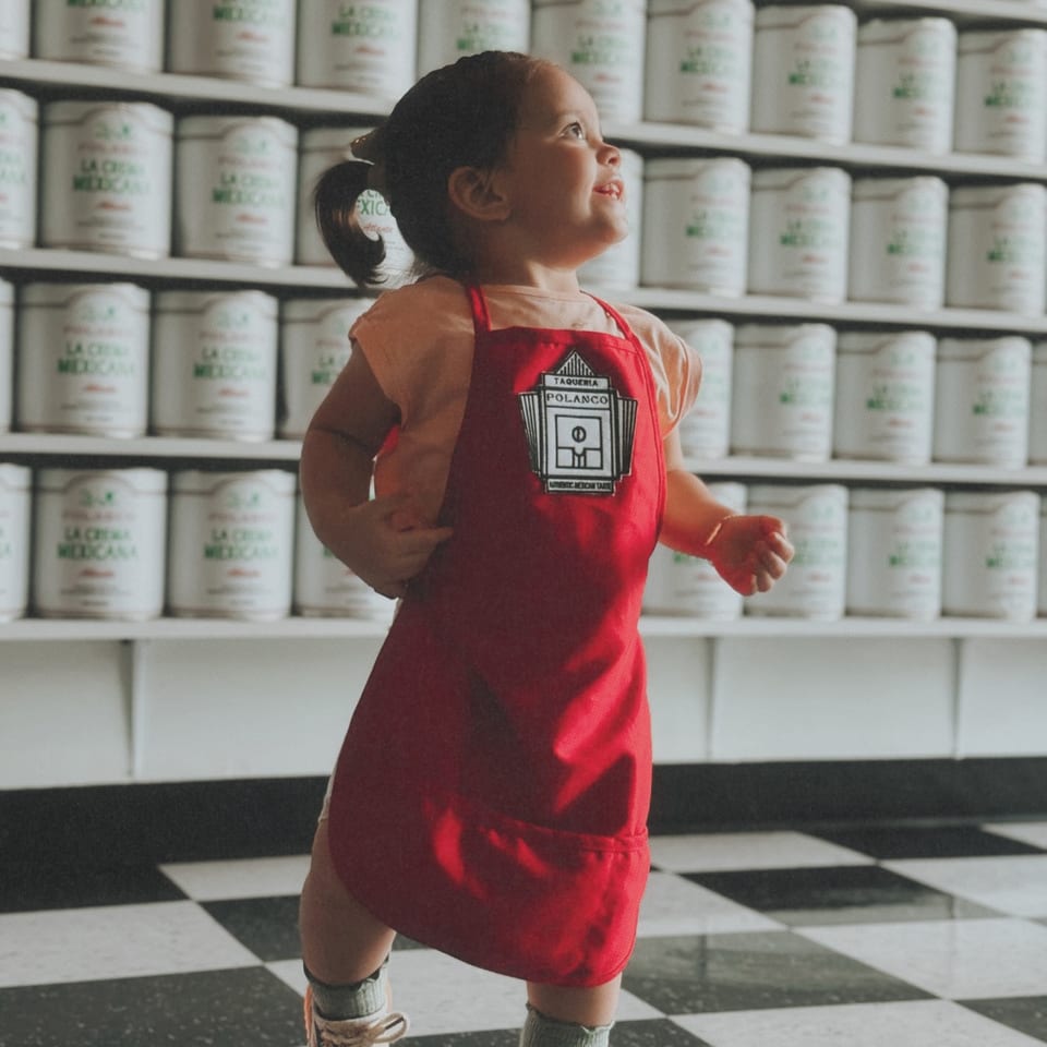 A young child in a red Polanco Taqueria apron stands on a checkered floor, looking up.