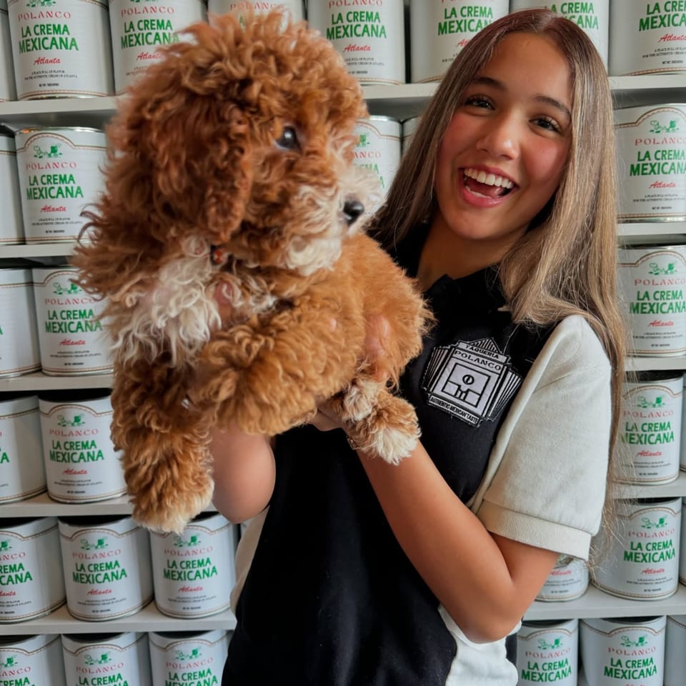 A smiling woman in a Polanco Taqueria shirt holds a fluffy brown puppy in front of shelves of "La Crema Mexicana" cans.