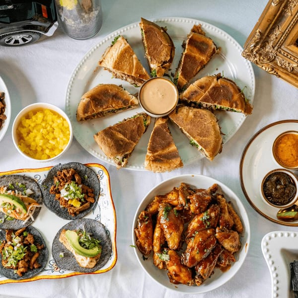 Overhead view of a Mexican feast with tacos, wings, torta slices, and various sauces on a white tablecloth.