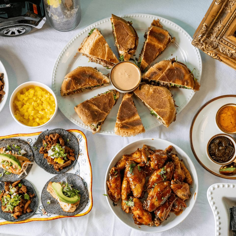 Overhead view of a Mexican feast with tacos, wings, torta slices, and various sauces on a white tablecloth.